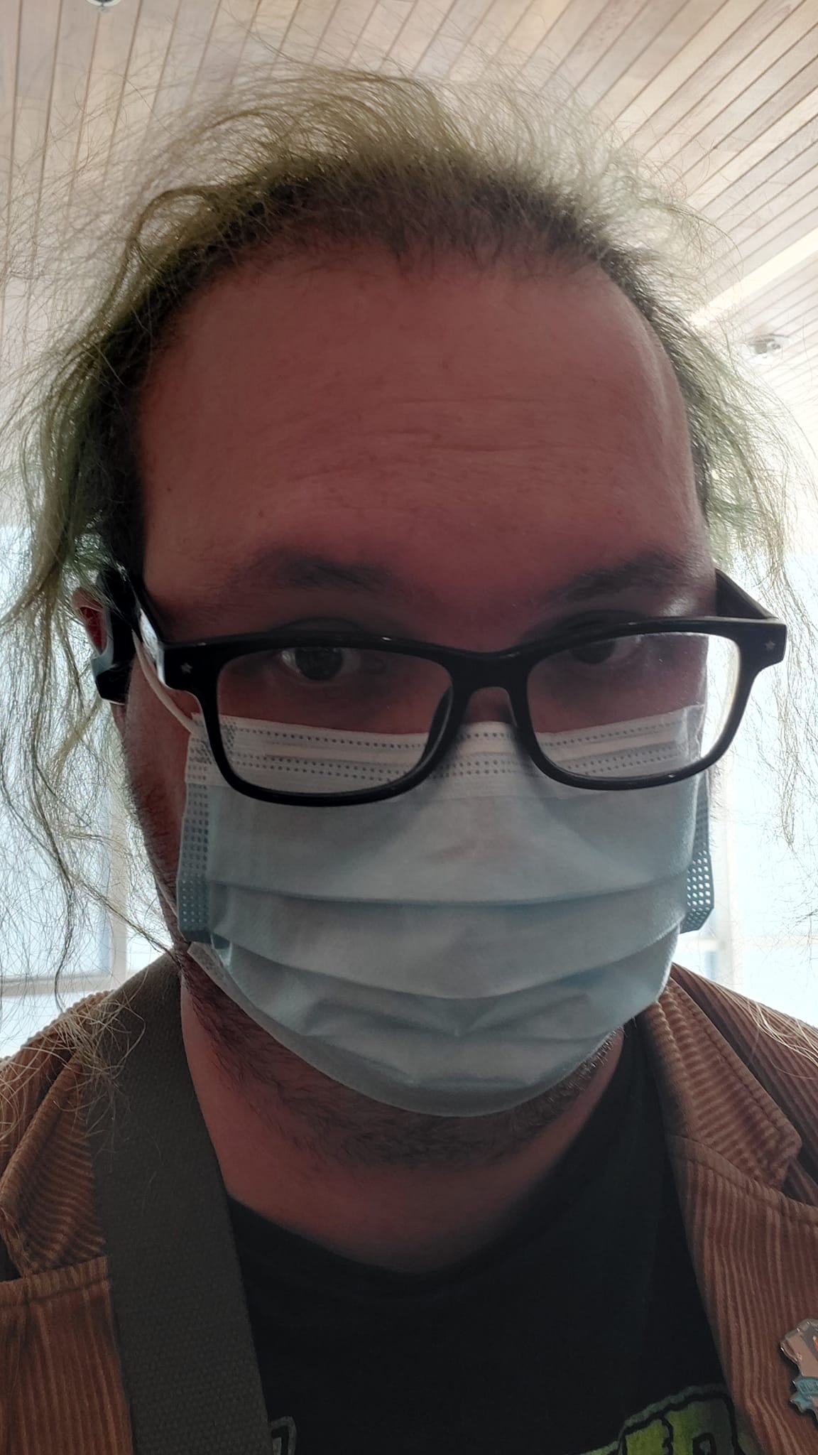 A man with green hair wearing a brown corderoy jacket sits in a hospital waiting room wearing a surgical mask